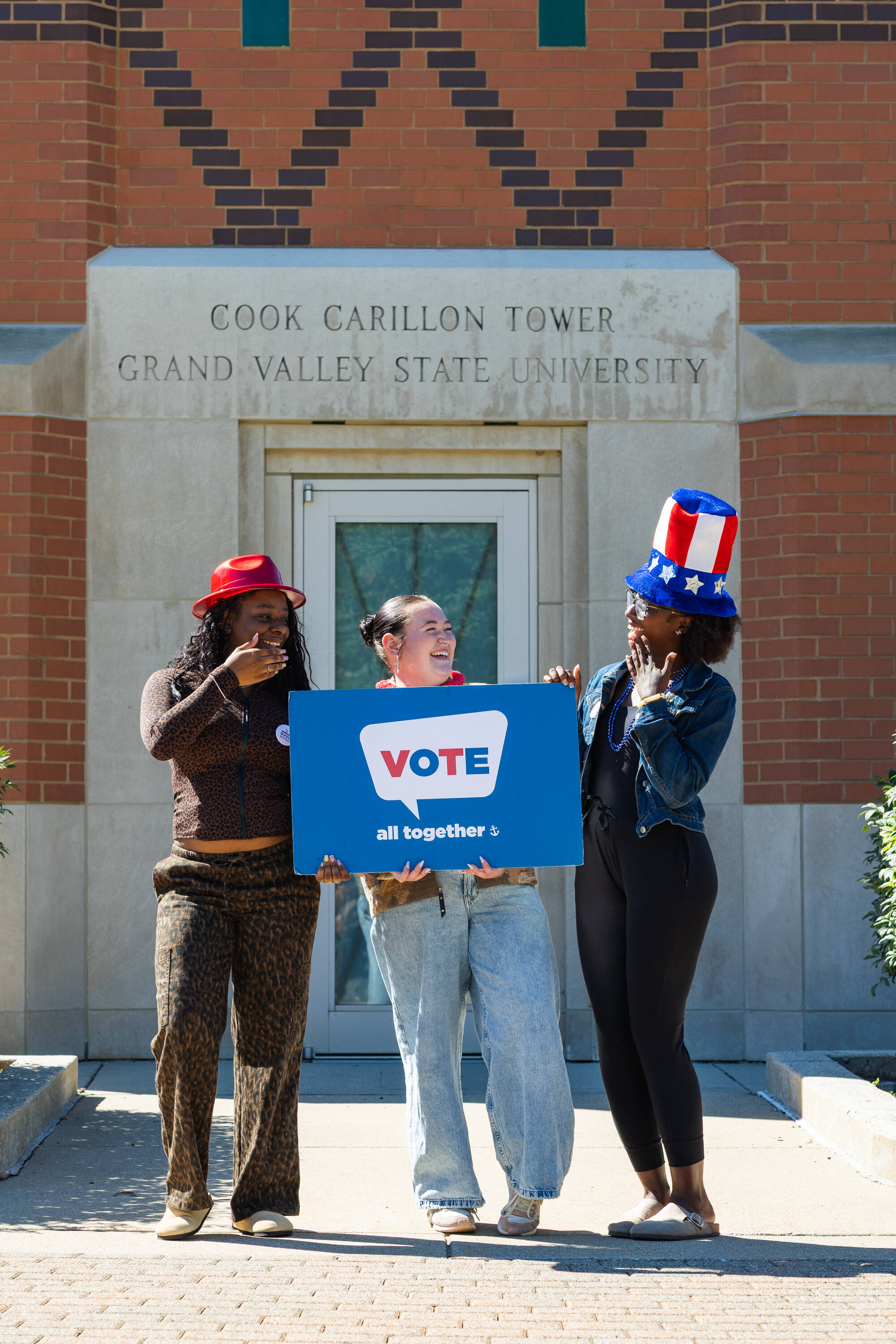 Three students laughing and holding a sign that says "vote all together"